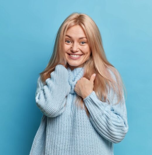 Glad blonde European female model looks gently at camera feels satisfied smiles toothily enjoys wearing new knitted sweater poses against blue background. Good emotions and feelings concept.