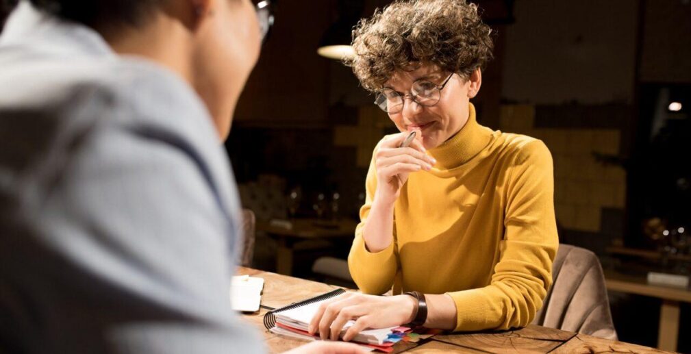 Content confident smart business lady in glasses sitting at table and holding diary, she smirking while talking to colleague in dark office