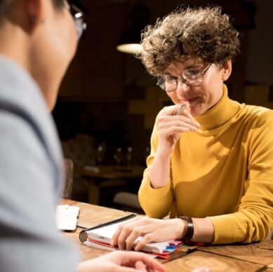 Content confident smart business lady in glasses sitting at table and holding diary, she smirking while talking to colleague in dark office